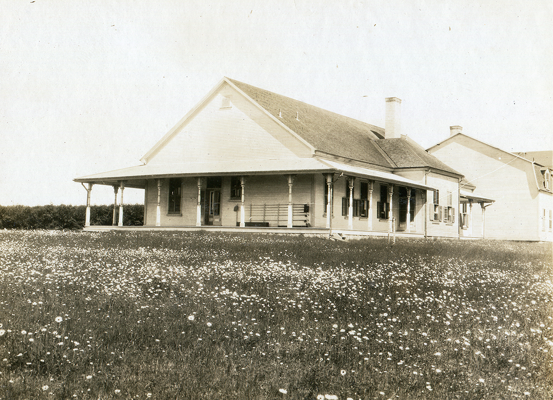 Photographie noir et blanc des façades nord et ouest de villa Estevan. L’extérieur du bâtiment est en déclin de bois et la toiture à double versant est faite de bardeau de cèdre. Les fenêtres sont munies de volets Le camp de pêche est muni d’une généreuse véranda sur laquelle est placée le support à canne a pêche. Au bout du bâtiment principal est raccordé au bâtiment secondaire occupé par les domestiques du propriétaire. Le bâtiment à 2 étages est légèrement reculé vers l’ouest par rapport au bâtiment principal. Le bâtiment est entouré d’un pré fleuri. L’on aperçoit l’immense haie de cèdre qui occupe l’espace avant du camp de pêche. Hormis sa taille, le camp de pêche est assez sobre, il n’y a que les colonnes du toit de la véranda qui souligne le style victorien de l’architecture du bâtiment.