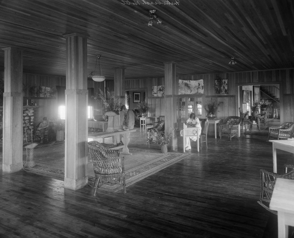 Photographie noir et blanc de l’intérieur du vaste hall de l’hôtel Boule Rock. Entièrement fait de bois, le hall est lumineux et bien rangé. Au centre de l’espace, un tapis recouvre le plancher de bois et l’on devine une portion du manteau de cheminée en pierres des champs. Des fauteuils en rotin et des tables sont répartis à travers la pièce.  On y retrouve également des bouquets de fleurs sur la majorité des tables et sur le plancher. On y aperçoit également deux hommes et une femme. La femme est assise à un bureau de travail et semble écrire une lettre. L’un des hommes est assis sur une table console et semble lire. Le dernier homme est assis dans un fauteuil près de la cheminée et li son journal.