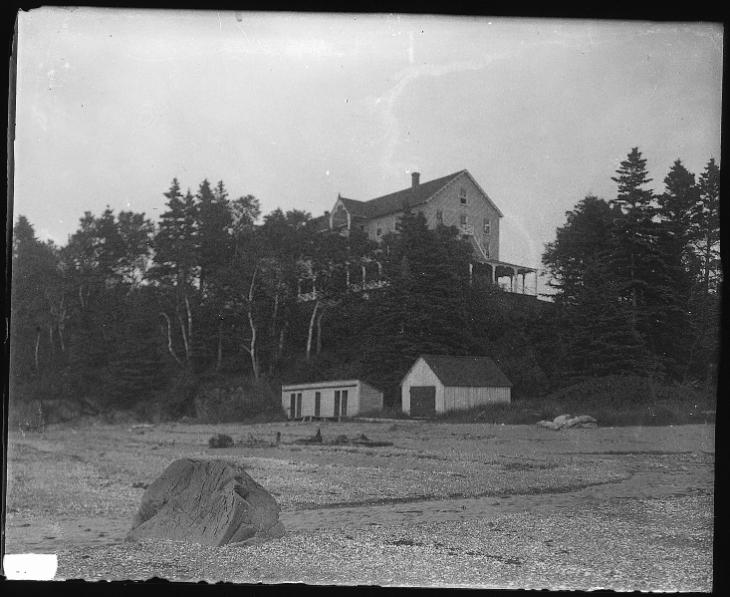 Photographie en noir et blanc de l’hôtel Cascade. Cet hôtel en bois de trois étages est situé au sommet d’une falaise escarpée. La falaise est couverte d’une forêt composée d’épinettes et de bouleaux blancs. Au pied de la falaise se trouve la plage rocheuse de Métis. Entre le bas de la falaise et le début de la plage, on aperçoit un petit hangar à bateaux et des cabines de bain, tous deux construits en bois.
