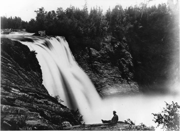 Photographie en noir et blanc de la chute sur la rivière Métis. La cascade de 90 pieds est bordée de deux falaises rocheuses abruptes entourées de forêt. Le photographe a capté la chute depuis le sommet de la falaise située à gauche. À quelques mètres du point de prise de vue, un homme est assis, les jambes étendues au bord de la falaise. Il observe la chute et le courant rapide de la rivière. Le photographe a utilisé une longue exposition, ce qui donne à l’eau de la rivière, s’écoulant du sommet de la chute, l’apparence d’un large filet blanc.