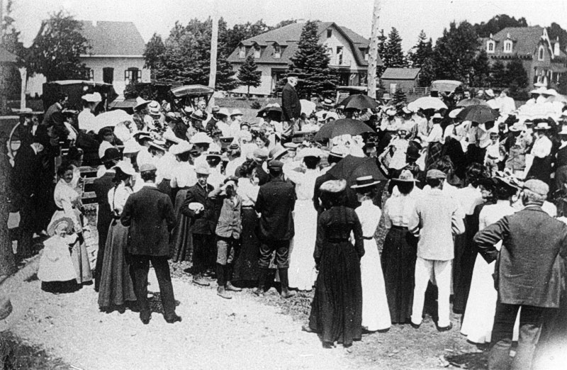 Photographie noir et blanc d'un regroupement de personnes lors de la célébration du Jubilée de la reine Victoria en 1897. Les gens sont regroupés à l'extérieur sous le soleil du midi. Au centre de la scène, un homme se tient debout et surplonbe la foule qui l'entoure. On apreçoit en arrière plan quelques sombtueuses résidences de villégiature de Métis-sur-Mer.
