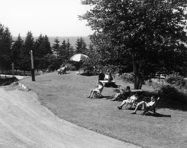 Photographie noir et blanc de vacanciers assis dans des chaises de jardin. Certains sont au soleil, d’autres sont à l’ombre sous un arbre ou sous un parasol. Ils sont situés sur un parterre aménagé. Devant eux passe une route de terre. Possiblement l’entrée de leur hôtel.   En arrière-plan apparaît, derrière une rangée d’épinettes, le fleuve Saint-Laurent.