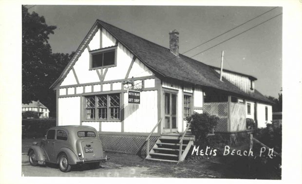 Photographie noir et blanc de la façade nord et ouest du «Metis Beach Gift Shop». La boutique souvenir est une résidence de deux étages blanche avec une toiture en bardeau de cèdre.  Sous la fenêtre de la façade nord est stationnée une voiture. Sur cette même façade est suspendu l’insigne de la boutique.