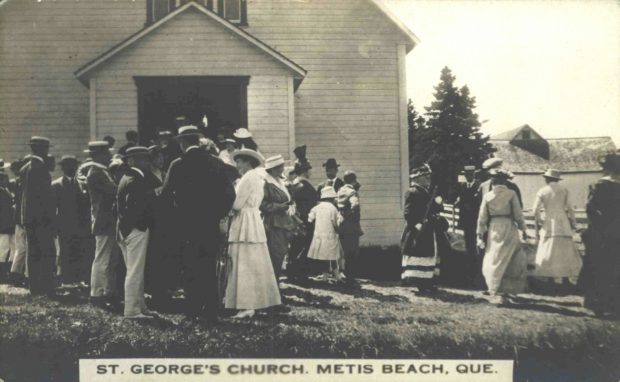 Photographie noir et blanc d'hommes et de femmes attroupés devant un église de bois à la fin de la messe. Les hommes sont, en majorité, vêtu d'un habit au veston foncé, d'un pentalon blanc et d'un chapeau tressé plat décoré d'un ruban. Les femmes portent une longue robe blanche et un chapeau plat tressé.  En arrière plan, on aperçoit derrière un massif d'épinettes matures, un bâtiment de ferme. 
