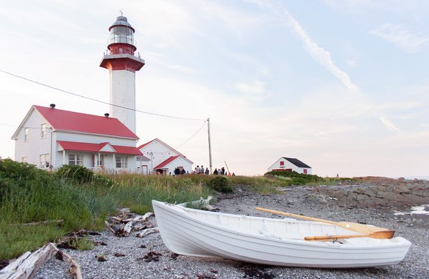 Photographie couleur récente du Phare de Métis-sur-Mer lors fin de journée d’été. En avant-plan une chaloupe blanche repose sur la berge. En arrière-plan, on aperçoit la maison du gardien, le phare, le bâtiment de la corne de brume et un petit hangar. Tous les bâtiments sont blancs et on un toit rouge. Une petite foule est rassemblée près des bâtiments. 