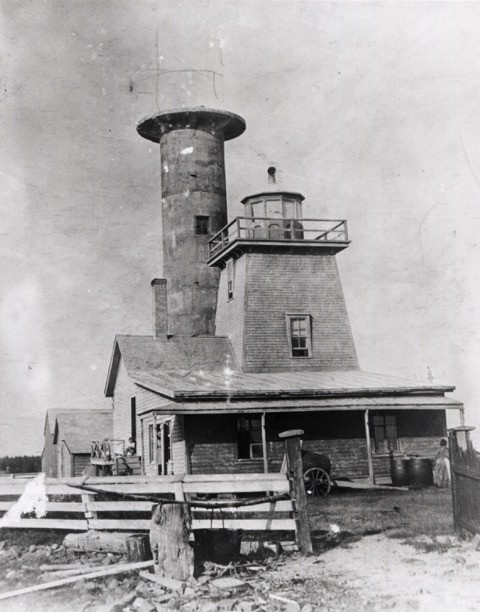 Photographie noir et blanc de la façade du premier phare de bois. Derrière le premier phare apparait la tour de ciment du nouveau phare qui est en construction. Il ne manque que la coupole vitrée qui devra accueillir la lanterne. Cette portion a été esquissée à la main au crayon de plomb sur la photographie.