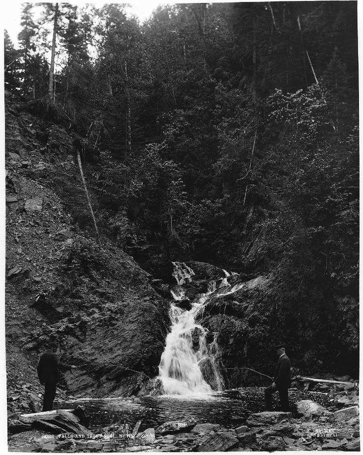 Photographie en noir et blanc de deux hommes en costume pêchant la truite au pied d’une chute d’eau. Les deux rives du ruisseau sont rocheuses. À l’arrière-plan, une falaise escarpée partiellement couverte par une forêt.