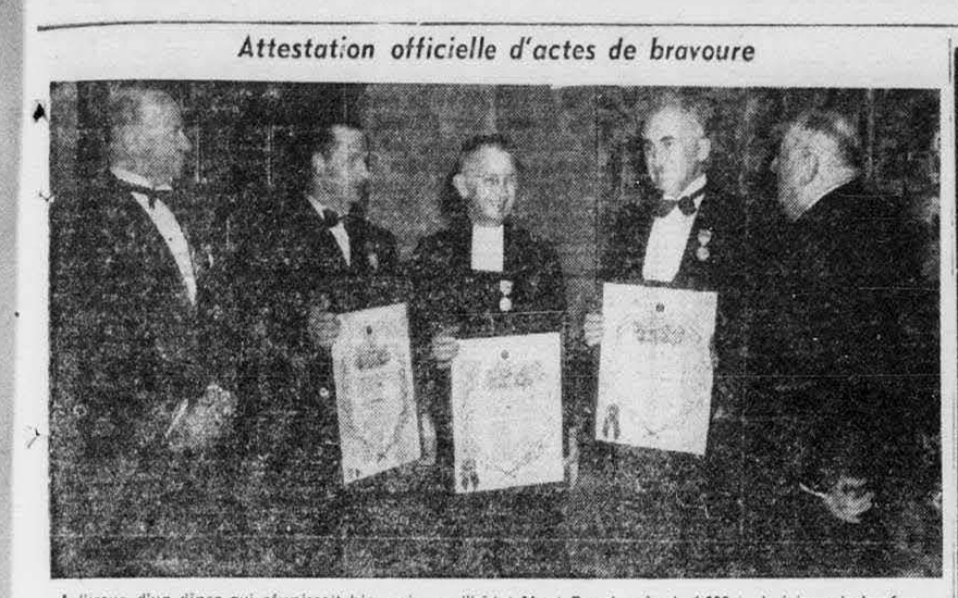 Photographie en noir et blanc tirée d’un journal, montrant cinq hommes vêtus de costumes noirs. Les trois hommes au centre tiennent des certificats blancs. Les deux hommes placés aux extrémités ne portent rien dans les mains.
