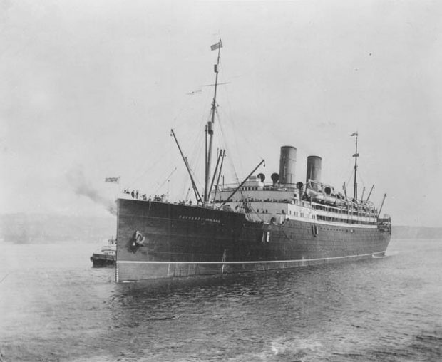 Photographie en noir et blanc du S.S. Empress of Ireland. Le navire est doté de deux cheminées. Des passagers sont visibles sur le pont avant. Le pavillon britannique (Union Jack) est hissé à la proue du navire.