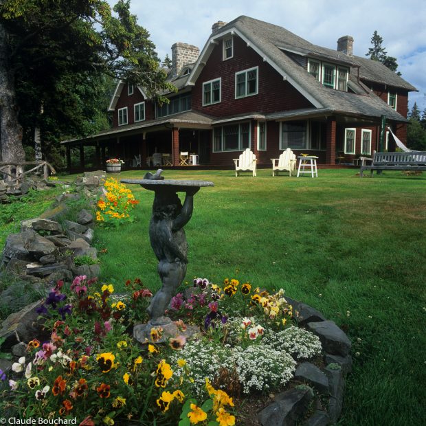 Photographie couleur de la façade d’une résidence d’été située à Métis-sur-Mer.  Au premier plan, une sculpture représentant un chérubin portant un bain d’oiseaux sur sa tête. La sculpture est placée au centre d’un parterre de pensées aux couleurs jaune, orange, blanche, mauve et rose.  La maison est recouverte de bardeaux de cèdre couleur bourgogne. Cette résidence à deux étages possède un toit en bardeaux de cèdre naturel et une véranda couverte à l’avant. Deux chaises Adirondack blanches sont disposées côte à côte sur la pelouse.