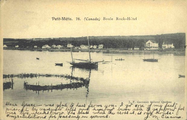 Carte postale de Metis montrant le village depuis le fleuve. Au premier plan, une installation de pêche à la fascine. Plusieurs petites embarcations de plaisance sont visibles sur l’eau.