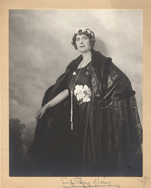 Photographie en studio en noir et blanc de Lady Byng. Lady Byng pose devant un décor peint représentant des nuages et des arbres. Elle porte une cape noire en satin, dentelle et fourrure, ainsi qu’une robe noire ornée de deux fleurs blanches en tissu à la ceinture. Elle est coiffée d’une couronne faite de feuilles d’érable. Sa main gauche repose sur sa hanche, permettant d’apprécier les détails de sa cape et de sa robe. Son bras droit est tendu et elle tient son manteau pour le laisser ouvert. La photographie est montée sur une carte signée de sa main « Evelyn Byng of Vimy » et du photographe « Rice ».