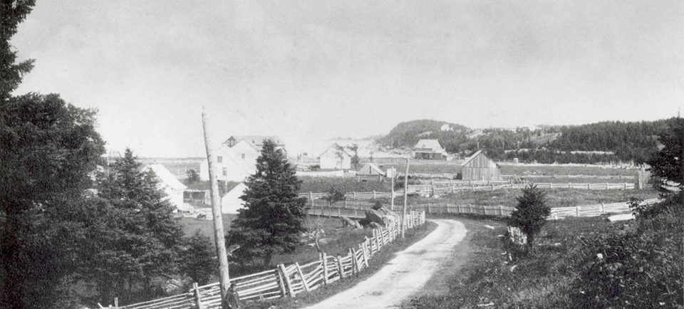 Photographie en noir et blanc d’un chemin de terre serpentant entre des maisons, des champs et des granges. Une clôture en bois longe le côté gauche du chemin. Un garçon est appuyé contre un poteau en bois.