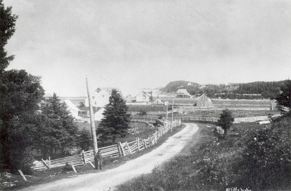 Photographie en noir et blanc d’un chemin de terre serpentant entre des maisons, des champs et des granges. Une clôture en bois longe le côté gauche du chemin. Un garçon est appuyé contre un poteau en bois.