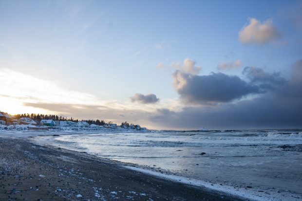 Photographie couleur du littoral de Métis en hiver, prise depuis la rive du fleuve Saint-Laurent.  La partie droite de l’image montre le fleuve à marée haute. Sur la gauche, on aperçoit les résidences bordant le rivage. La photo a été prise en fin d’après-midi. Une fine couche de neige recouvre les toits ainsi que la plage.