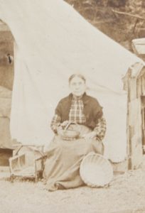 Image sépia d’une femme autochtone assise devant une tente, vendant des paniers tressés.