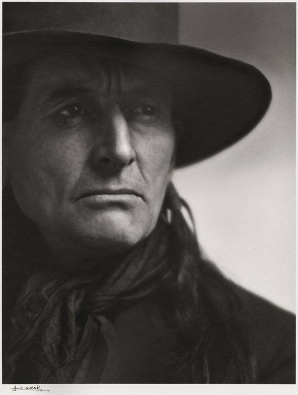 Portrait en noir et blanc de Grey Owl. Le portrait est un gros plan du visage de l’homme. Cadre à droite, le visage et le regard de Grey Owl sont légèrement tournés vers la droite. Vêtu de noir, il porte un chapeau de cowboy, un foulard et une veste. Une mèche de cheveux tombe sur son épaule. La photographie est signée Yousuf Karsh.