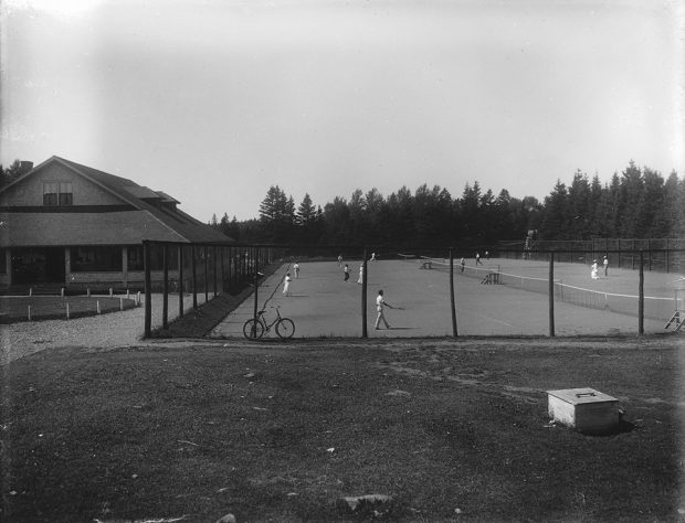 Photographie en noir et blanc des terrains de tennis du Cascade Golf and Tennis Club. Une haute clôture en bois entoure le court de tennis. Des hommes, vêtus de pantalons blancs et de chemises blanches, ainsi que des femmes, portant une longue jupe blanche, une chemise blanche à manches longues et un chapeau, jouent au tennis. Sur le côté gauche, on aperçoit le pavillon du club.