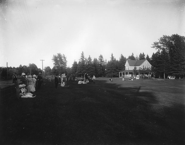 Photographie en noir et blanc du premier départ du Cascade Golf Club. Vingt personnes vêtues à la mode de 1914 observent et attendent. Certaines femmes sont assises sur l’herbe. À l’arrière-plan, un chalet de deux étages au style victorien épuré.