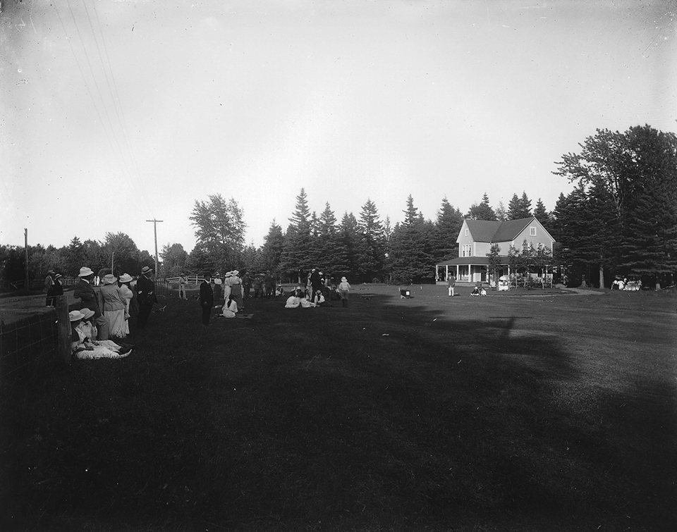 Photographie en noir et blanc du premier départ du Cascade Golf Club. Vingt personnes vêtues à la mode de 1914 observent et attendent. Certaines femmes sont assises sur l’herbe. À l’arrière-plan, un chalet de deux étages au style victorien épuré.