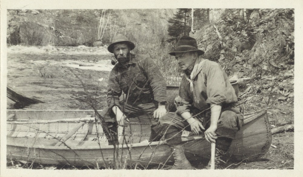 Les prospecteurs sont assis sur le bord de leur canot sur la rive. À l’arrière-plan, on aperçoit les rapides d’une rivière et un paysage rocheux bordé d’arbres.