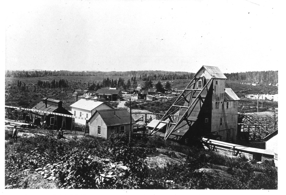 Photographie en noir et blanc du haut chevalement de la mine Tough-Oakes que l’on voit au premier plan avec d’autres bâtiments de la mine. De petites maisons bordent une route qui serpente dans une grande clairière. Une forêt profonde se trouve à l’arrière-plan.