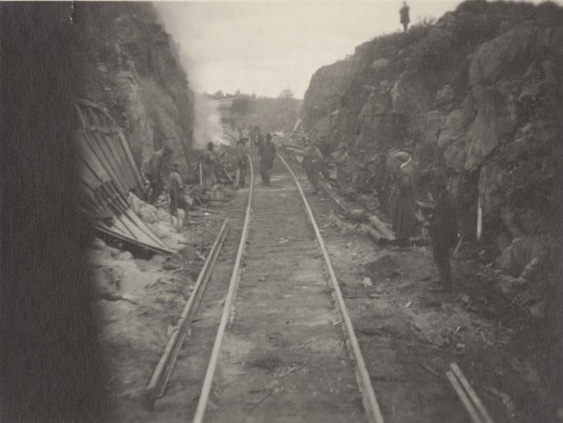 Photographie en noir et blanc de la construction du Temiskaming and Northern Ontario Railway. La voie ferrée repose sur un terrain plat entre les roches dynamitées du bouclier cambrien. L’équipe du chemin de fer se tient à l’arrière-plan.