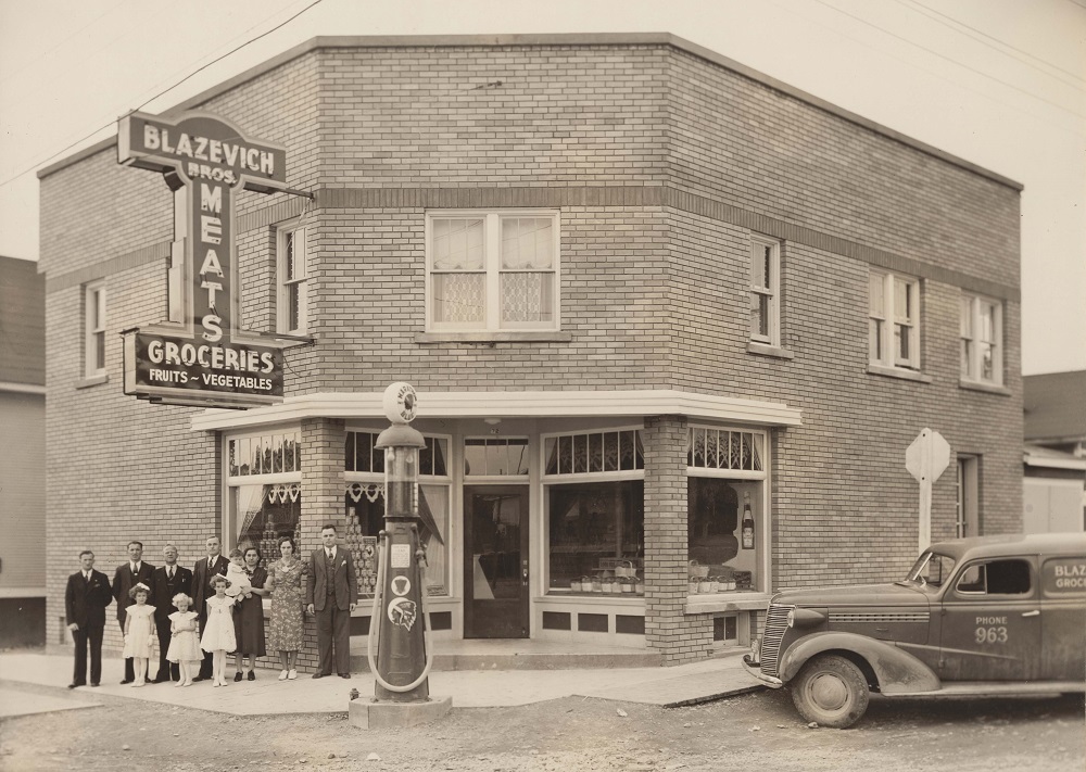 Photographie en noir et blanc de l’épicerie des frères Blazevich sur Main Street. La famille Blazevich est debout sous l’enseigne au néon fixée au bâtiment en brique. Une pompe à essence à l’ancienne est au premier plan, et le véhicule de livraison des frères Blazevich se trouve à droite de l’image.