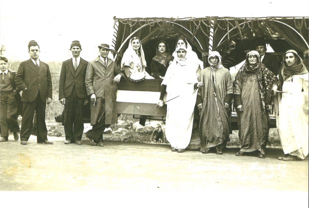 Photographie en noir et blanc de personnes vêtues de vêtements traditionnels syriens avec un char allégorique du jour du couronnement. Trois femmes et un homme sont assis sur le char allégorique, tandis que quatre hommes sont debout par-devant. Trois hommes en costume et un garçon se tiennent à gauche du char allégorique, l’un d’eux étant appuyé contre le char allégorique.