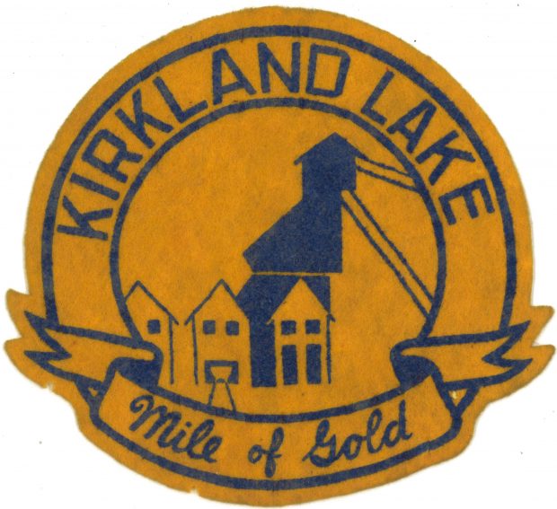 Un écusson en feutre de couleur or et bleu, avec les mots Kirkland Lake et Mile of Gold écrits autour de l’extérieur de l’écusson. L’image au centre de l’écusson représente des bâtiments de la mine et un chevalement au centre.