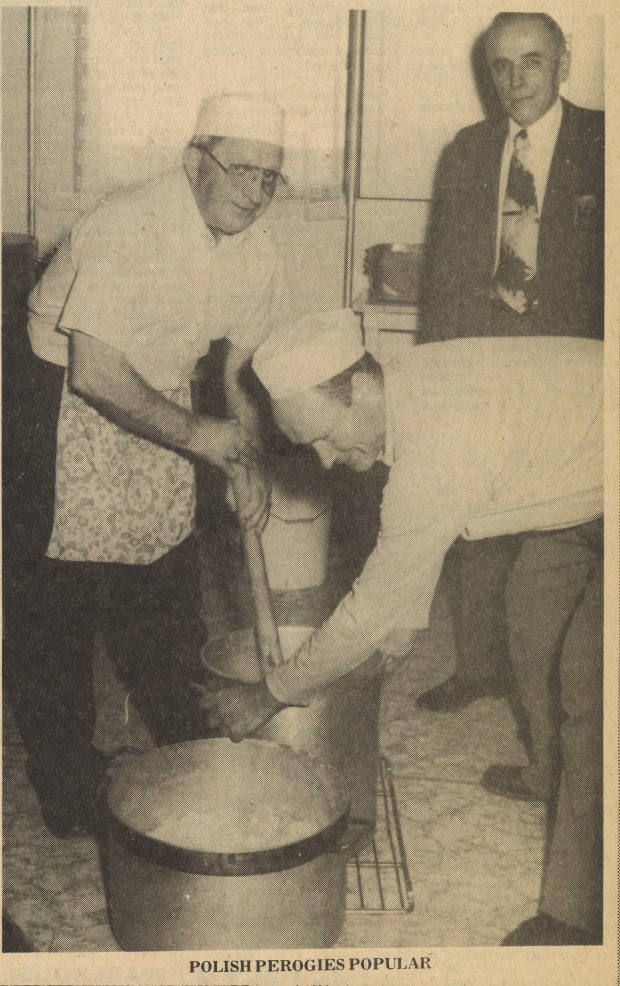 Coupure de presse d’une photographie – un homme portant un tablier court et tenant un grand bâton dans un grand pot en métal qu’un autre homme, habillé de la même façon, tourne sur le sol. Un homme en costume se tient à l’arrière-plan. La légende au bas de l’image est la suivante : « Pérogies polonais populaires ».