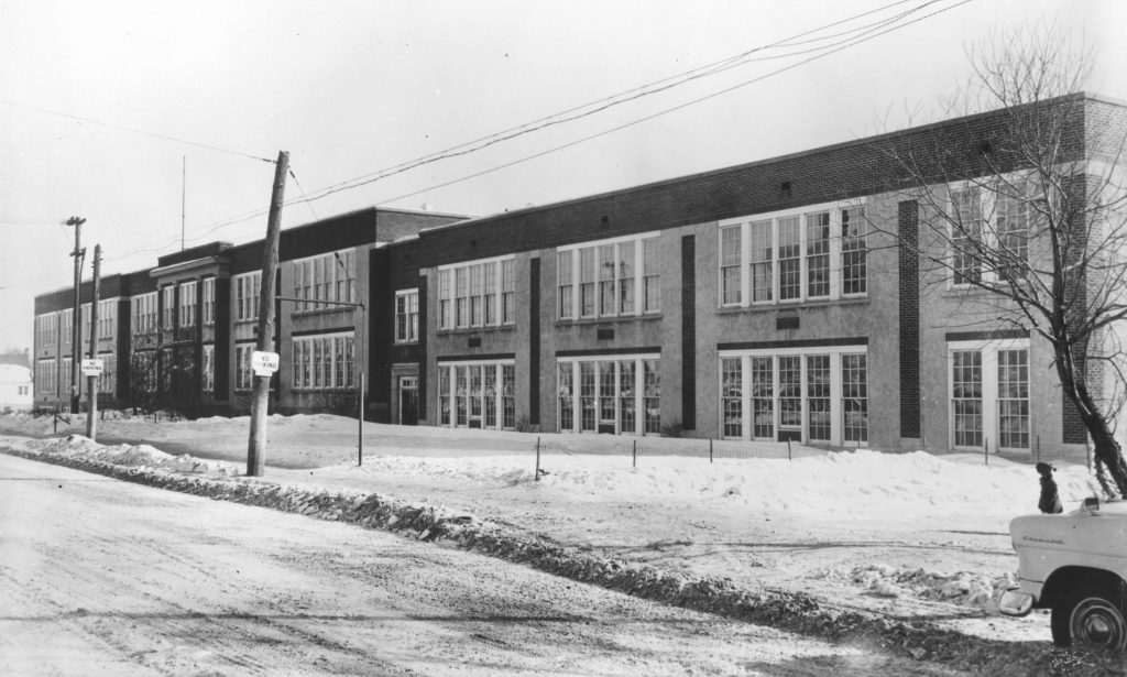 Photographie en noir et blanc d’une école secondaire, un bâtiment en brique de deux étages avec de nombreuses fenêtres. Il y a de la neige sur la pelouse avant. Deux poteaux téléphoniques se trouvent à gauche de la photo, l’avant d’une voiture blanche se trouve dans le coin inférieur droit.