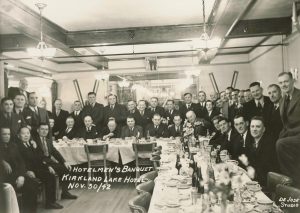 Photographie en noir et blanc du banquet des hôteliers à l’hôtel Kirkland Lake. Charlie Chow est assis, deuxième à partir de la gauche. Plus de 40 hommes en costume sont assis et debout autour des tables après un dîner, face à l’appareil photo. Les plateaux de table sont remplis de tasses de thé et de bouteilles de bière, de serviettes de table et d’assiettes vides. Quatre lustres sont suspendus au plafond.