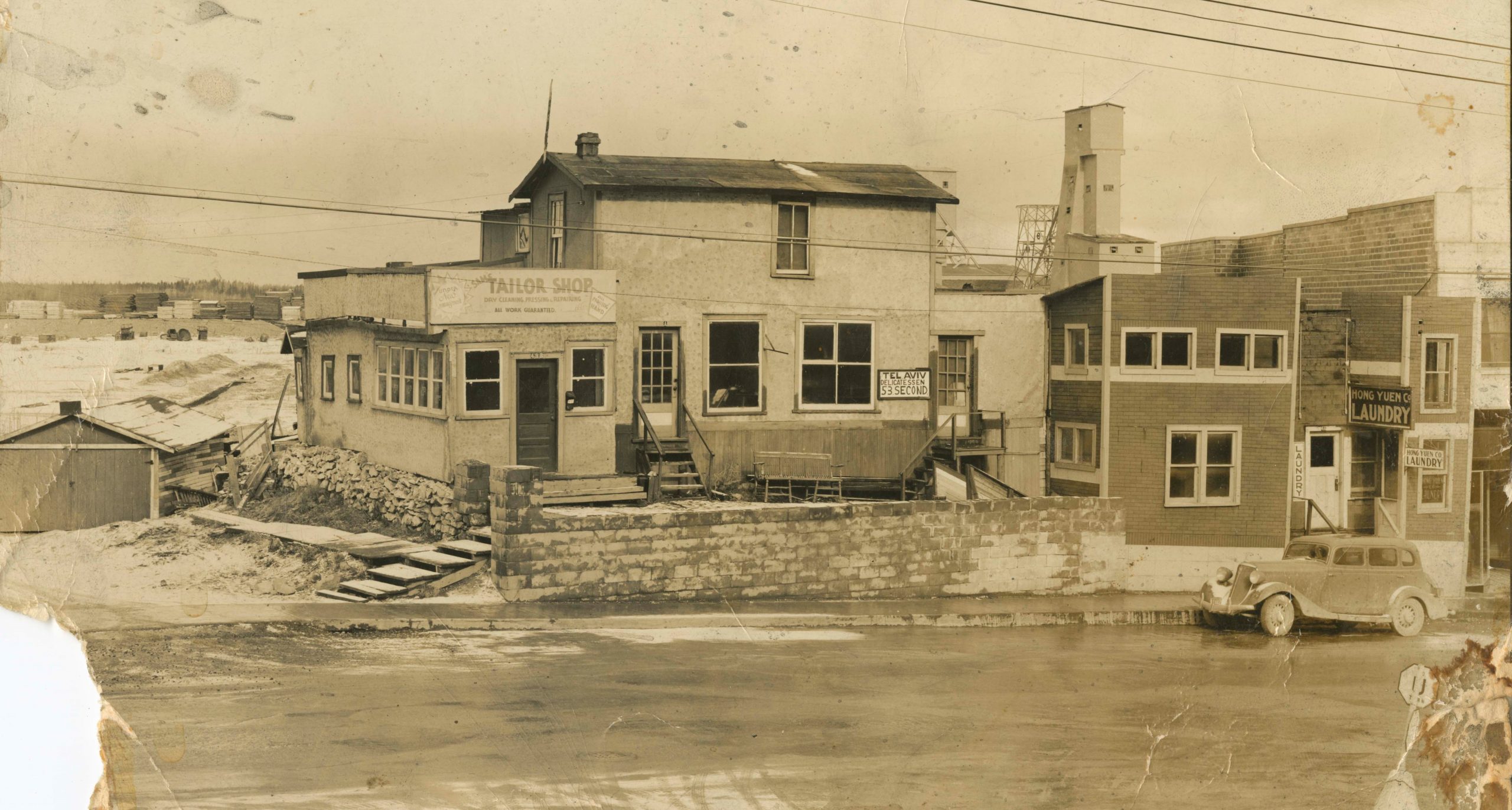 Photographie en noir et blanc d’une maison et de commerces sur une rue avec une voiture garée à droite de l’image. Les bâtiments de la mine sont à l’arrière-plan.