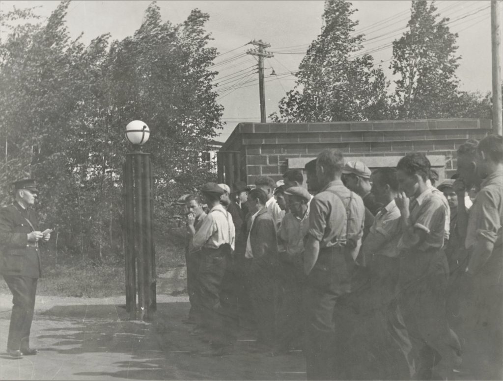 Photographie en noir et blanc d’un groupe d’hommes debout à côté d’un grand poteau de porte métallique, faisant face à un gardien en uniforme à l’entrée de la propriété de la mine. Le gardien tient une feuille de papier.