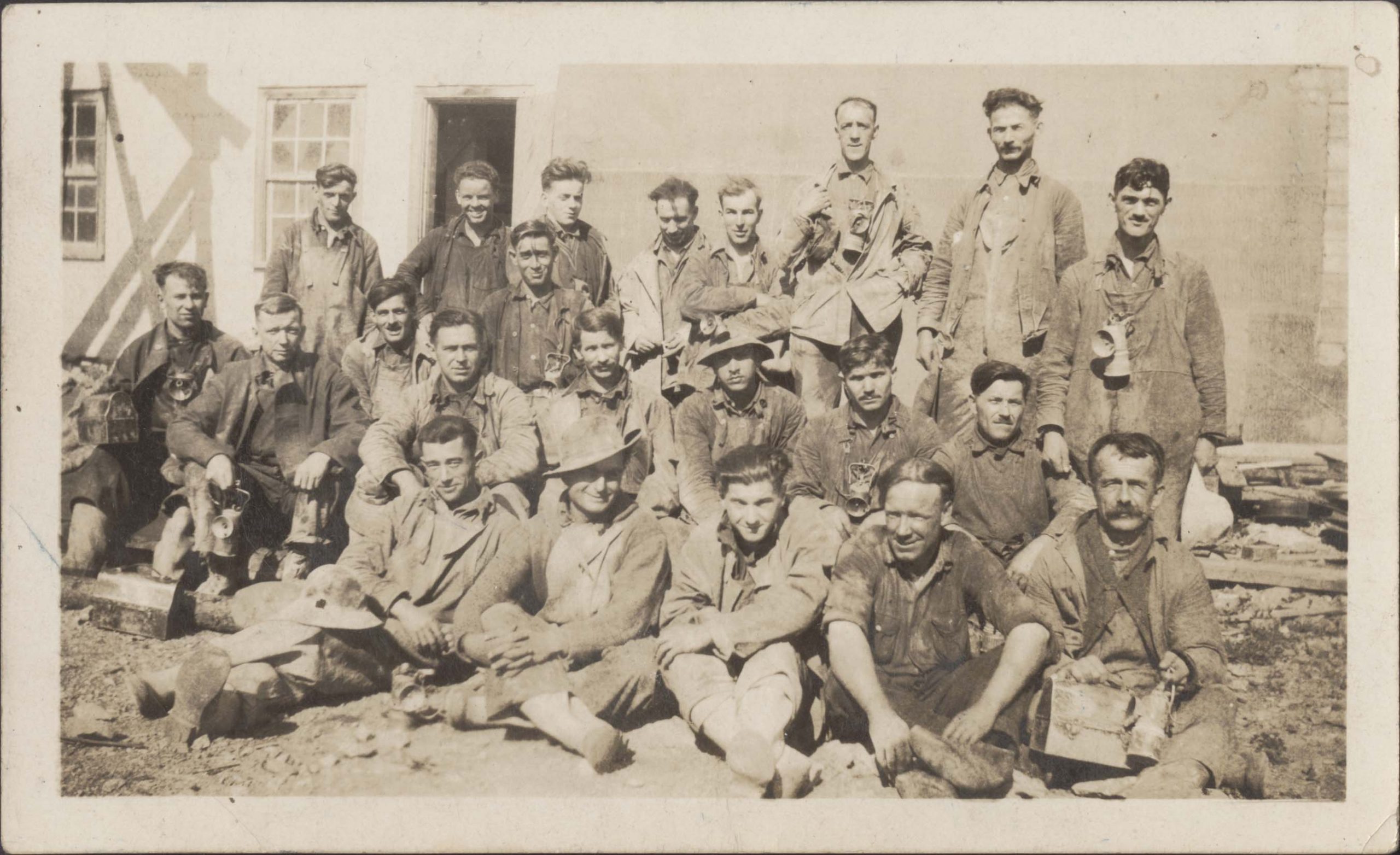 Photographie en noir et blanc de 22 mineurs portant des vêtements de travail sales : combinaisons, chemises, vestes, casques ou chapeaux. Certains hommes portent une lanterne autour du cou, et d’autres tiennent des boîtes à lunch. Des hommes sont assis et debout en arrière-plan, devant un bâtiment de la mine.