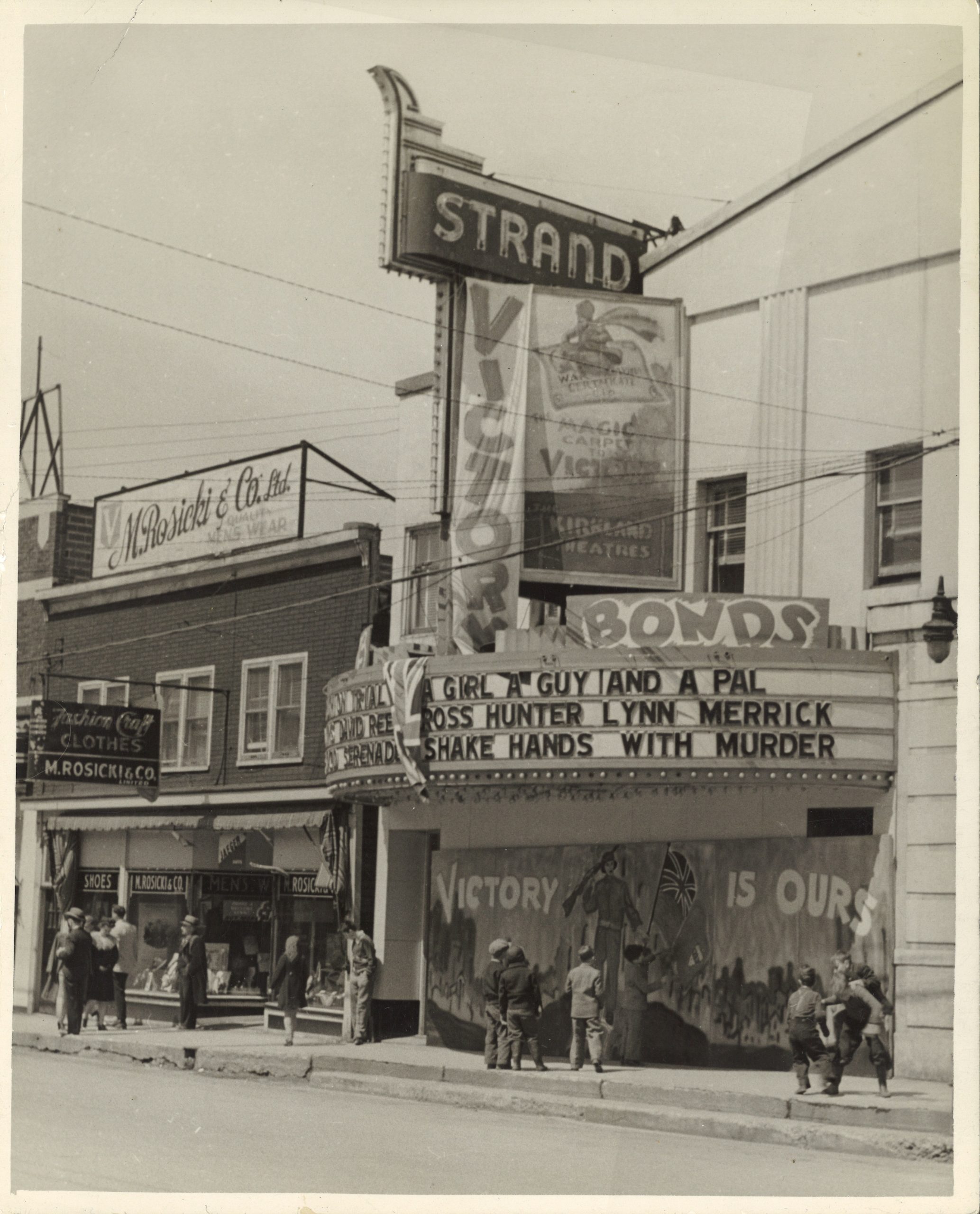 Photographie en noir et blanc de la façade du Strand Theatre. Le bâtiment est blanc et une grande marquise s’étend sur le trottoir devant. Des affiches de cinéma et des bannières d’obligations de la Victoire sont accrochées à la façade du bâtiment. Des hommes, des femmes et des enfants sont sur le trottoir. Un bâtiment en briques rouges avec des façades de magasins se trouve à gauche de la salle.