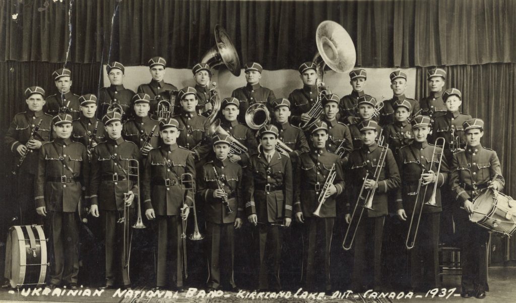 Photographie en noir et blanc de trois rangées d’hommes en uniformes tenant des instruments dans un studio de photographie. Les mots « Orchestre national ukrainien – Kirkland Lake, Ontario – Canada – 1937 » sont écrits en bas de la photo.