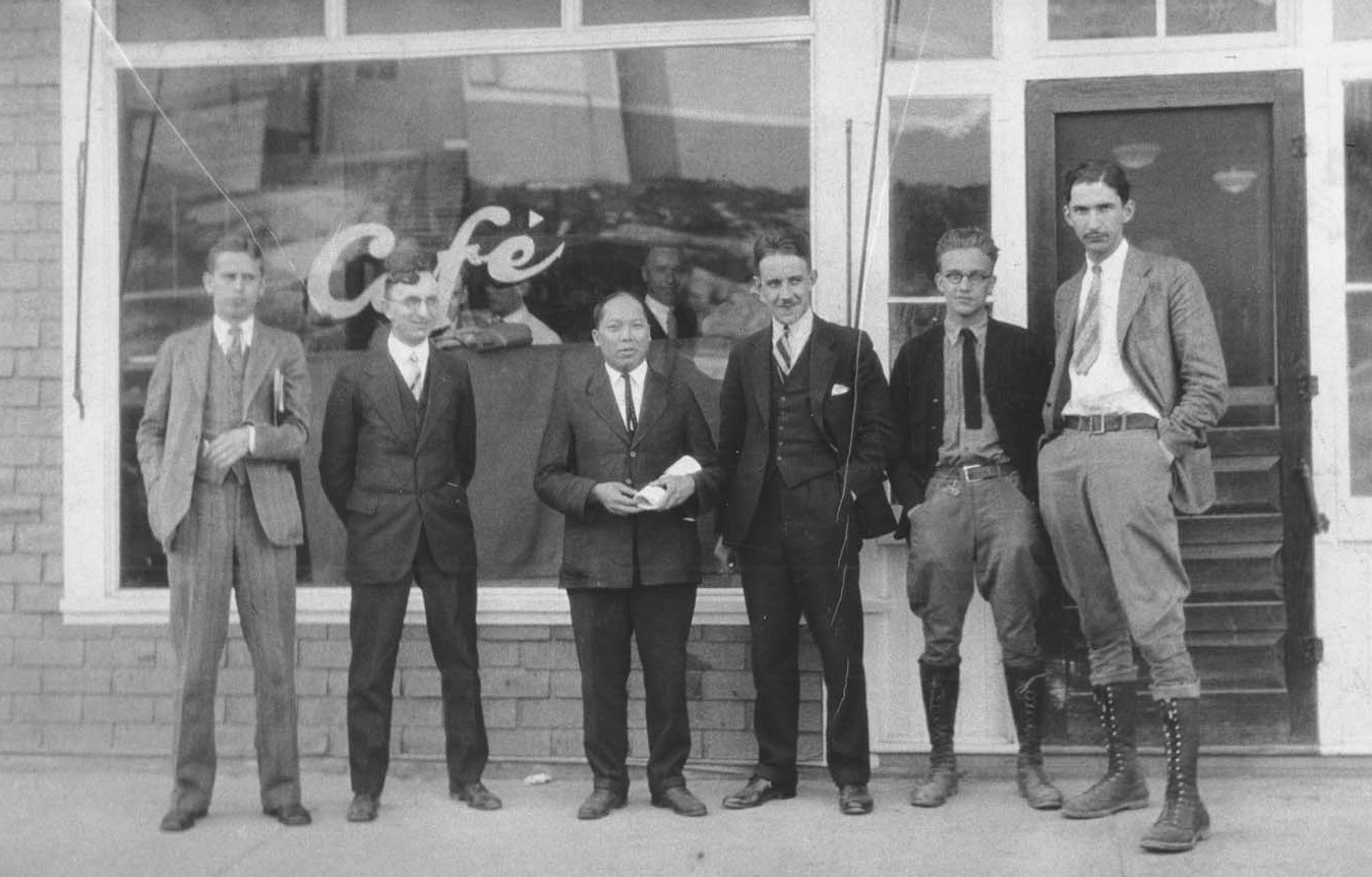 Photographie en noir et blanc de six hommes debout à l’extérieur d’un bâtiment dont la fenêtre avant porte l’inscription Café. Les hommes sont habillés en costume sur le trottoir.