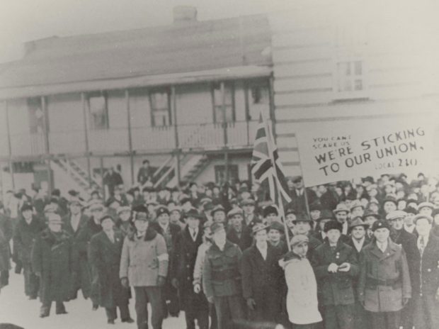 Photographie en noir et blanc d’une grande foule d’hommes vêtus de vêtements chauds se tenant à l’extérieur d’un grand bâtiment en hiver, tenant un drapeau britannique et une pancarte pro-syndicat.