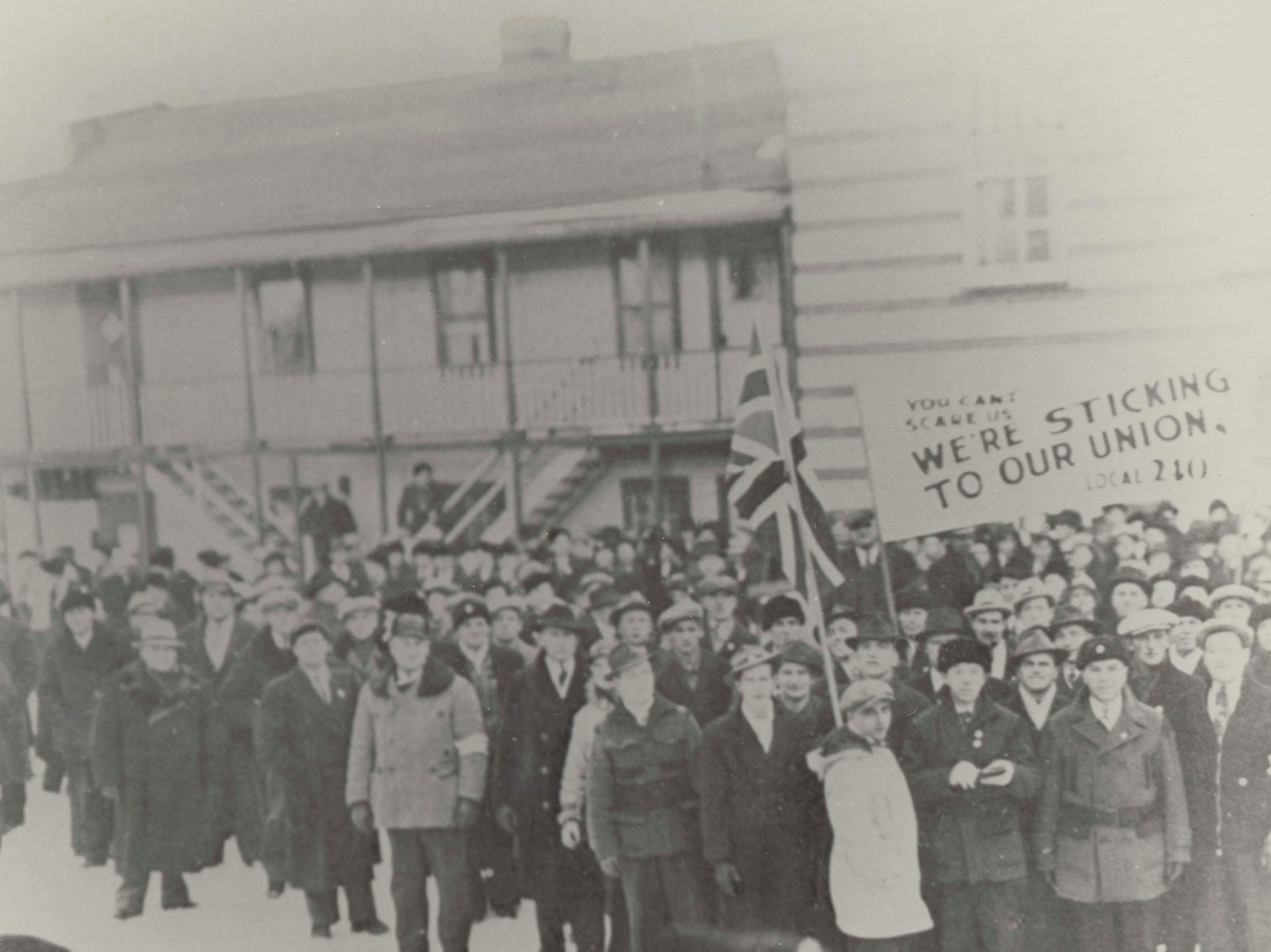Photographie en noir et blanc d’une grande foule d’hommes vêtus de vêtements chauds se tenant à l’extérieur d’un grand bâtiment en hiver, tenant un drapeau britannique et une pancarte pro-syndicat.
