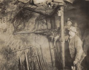 Photographie en noir et blanc de quatre hommes debout à côté d’une grande foreuse dans le tunnel souterrain d’une mine.