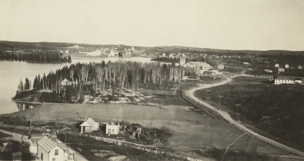 Photographie en noir et blanc prise depuis un point élevé, avec vue sur un terrain défriché, des mines et des maisons, avec une forêt au loin.