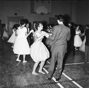 Photographie en noir et blanc de jeunes hommes en costume et de femmes en robe dansant dans un gymnase.