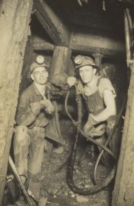 Photographie en noir et blanc de deux hommes travaillant sous terre dans une mine. Ils portent des casques de protection avec lampes et des vêtements de travail.