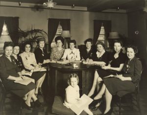 Photographie en noir et blanc de dix femmes assises autour d’une grande table ronde en bois. Une fille est assise sur un coussin devant la table et tient une feuille de papier.