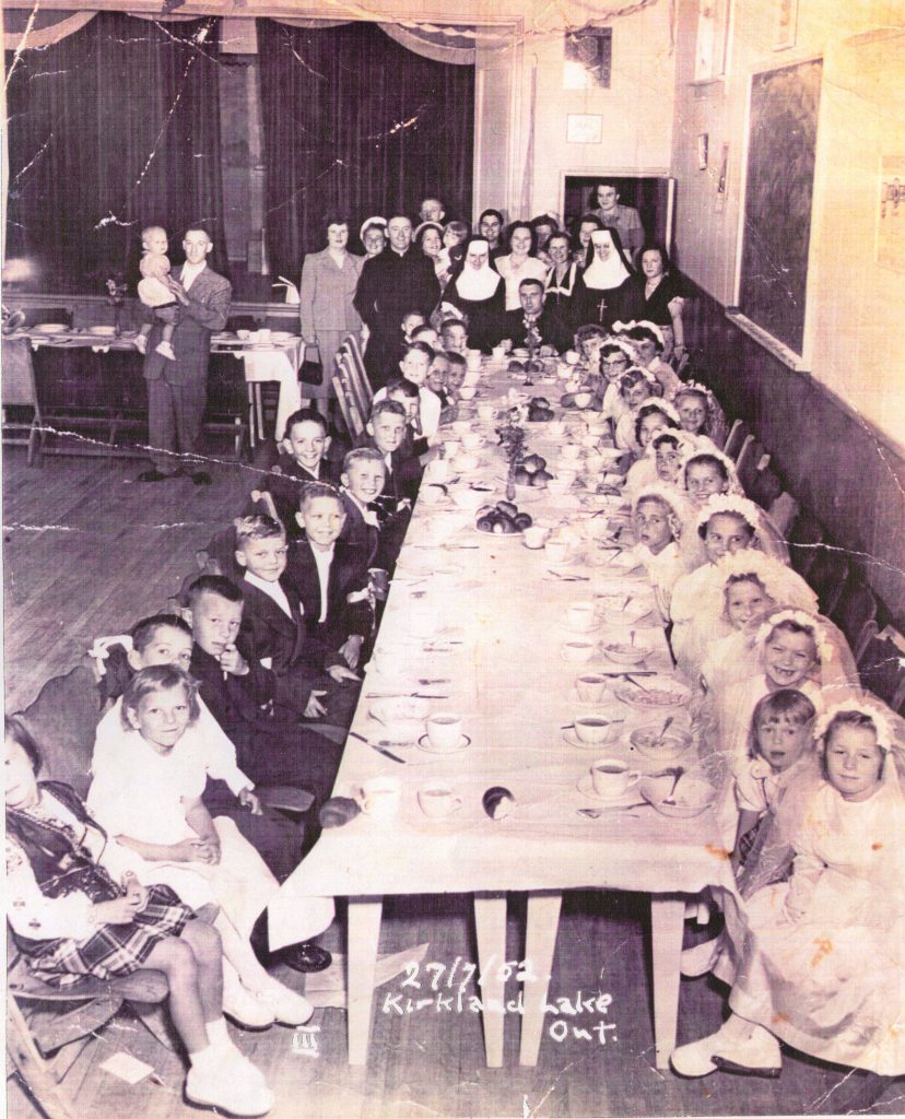 Photographie en noir et blanc d’environ 40 enfants assis autour d’une longue table en train de prendre un repas. Les garçons sont vêtus de costumes sombres, les filles portent des robes blanches pour la plupart. À l’arrière-plan de la salle se trouvent des hommes et des femmes, ainsi qu’un prêtre et deux religieuses.