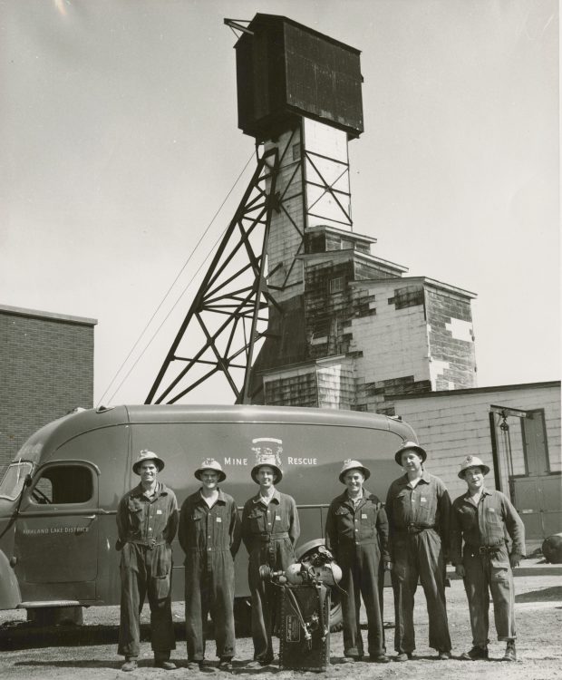 Photographie en noir et blanc de six hommes en salopette avec leur équipement devant eux. Ils sont debout devant leur camionnette. Un chevalement de mine est en arrière-plan.