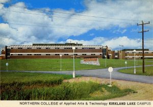 Carte postale en couleur d’un grand bâtiment de briques rouges à deux étages sur une colline avec un panneau sur la pelouse avant. L’enseigne indique Northern College of Applied Arts and Technology – Kirkland Lake Campus.