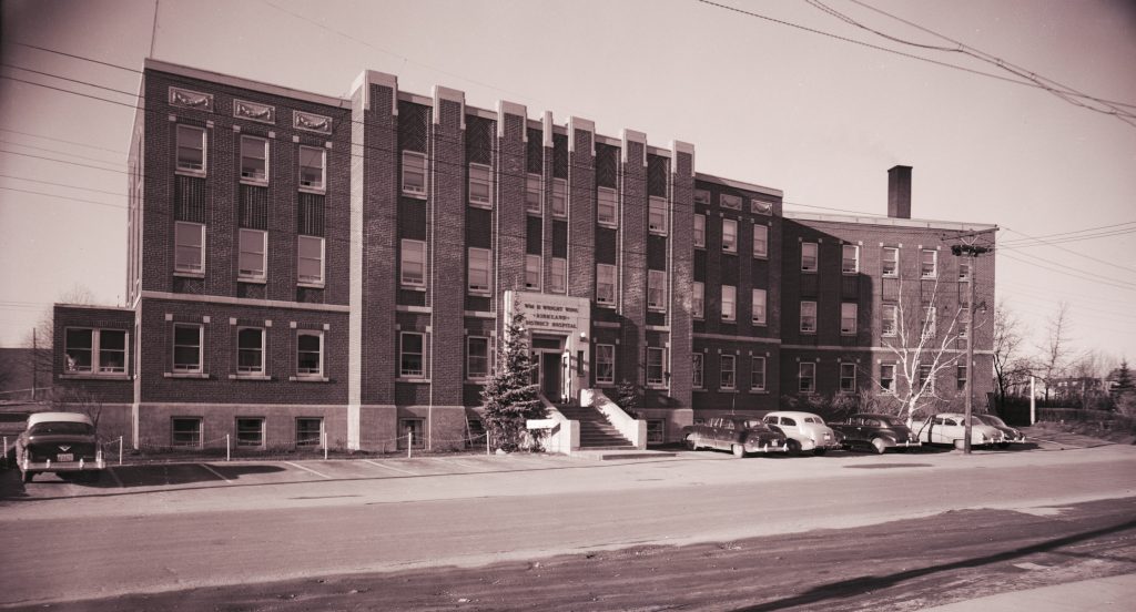 Photographie en noir et blanc de l’hôpital du district de Kirkland, un grand bâtiment en brique de quatre étages donnant sur une rue. Des voitures sont garées devant le bâtiment.